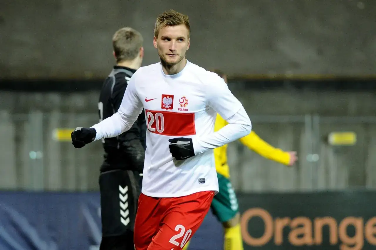 Piłkarz Kacper Przybyłko w białej koszulce z numerem 20 i czerwonych spodenkach, na tle innych zawodników.