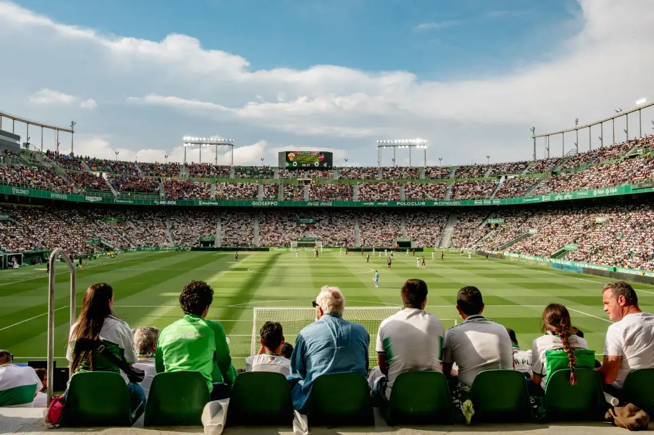Widok na pełny stadion podczas meczu Elche CF. Kibice na trybunach, piłkarze na murawie, a nad nimi niebo z chmurami.