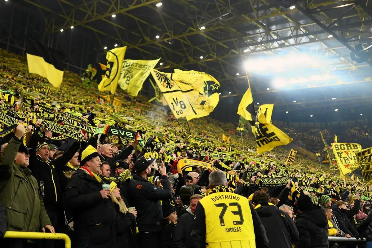 Tłum kibiców na stadionie, machający żółto-czarnymi flagami i szalikami. Atmosfera gorących rozgrywek w ramach bundesliga.