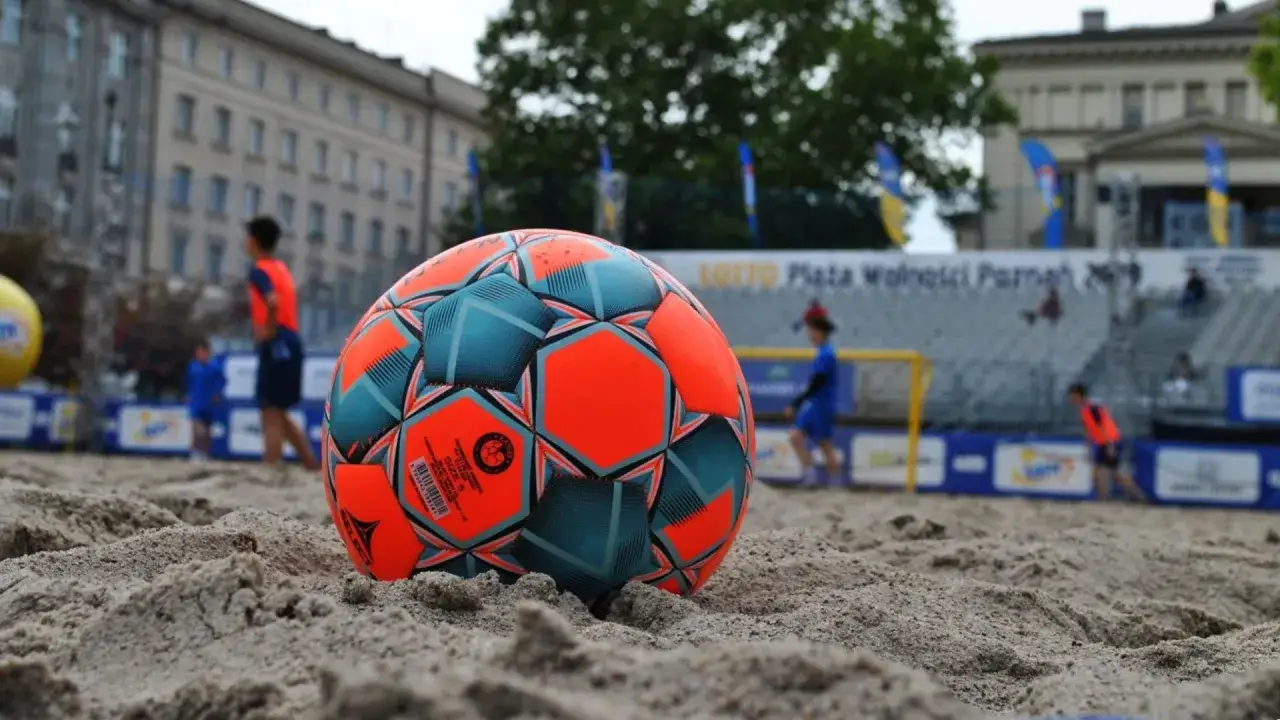 Piłka do beach soccer Polska leży na piasku. W tle widać boisko i zawodników.