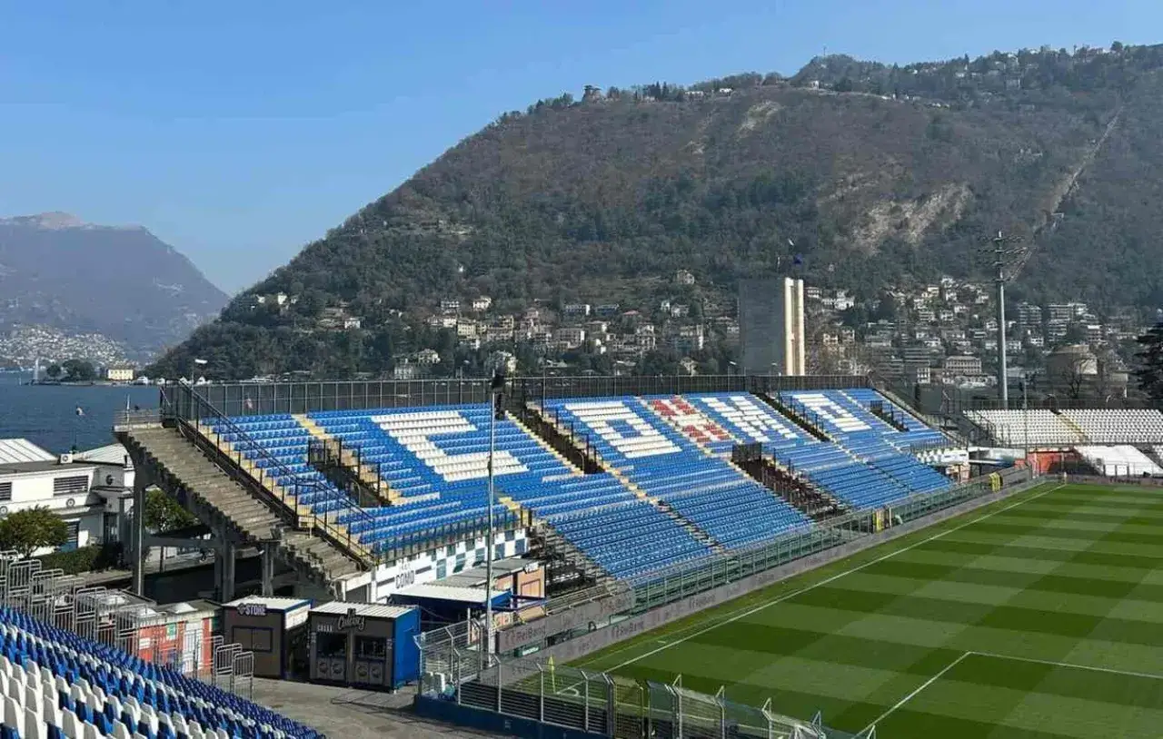 Stadion Como 1907 z niebieskimi i białymi siedzeniami, w tle jezioro i wzgórza z zabudowaniami.