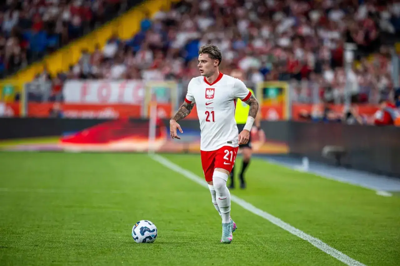 Piłkarz Nicola Zalewski w białej koszulce z numerem 21 i czerwonych spodenkach biegnie z piłką po zielonej murawie stadionu.
