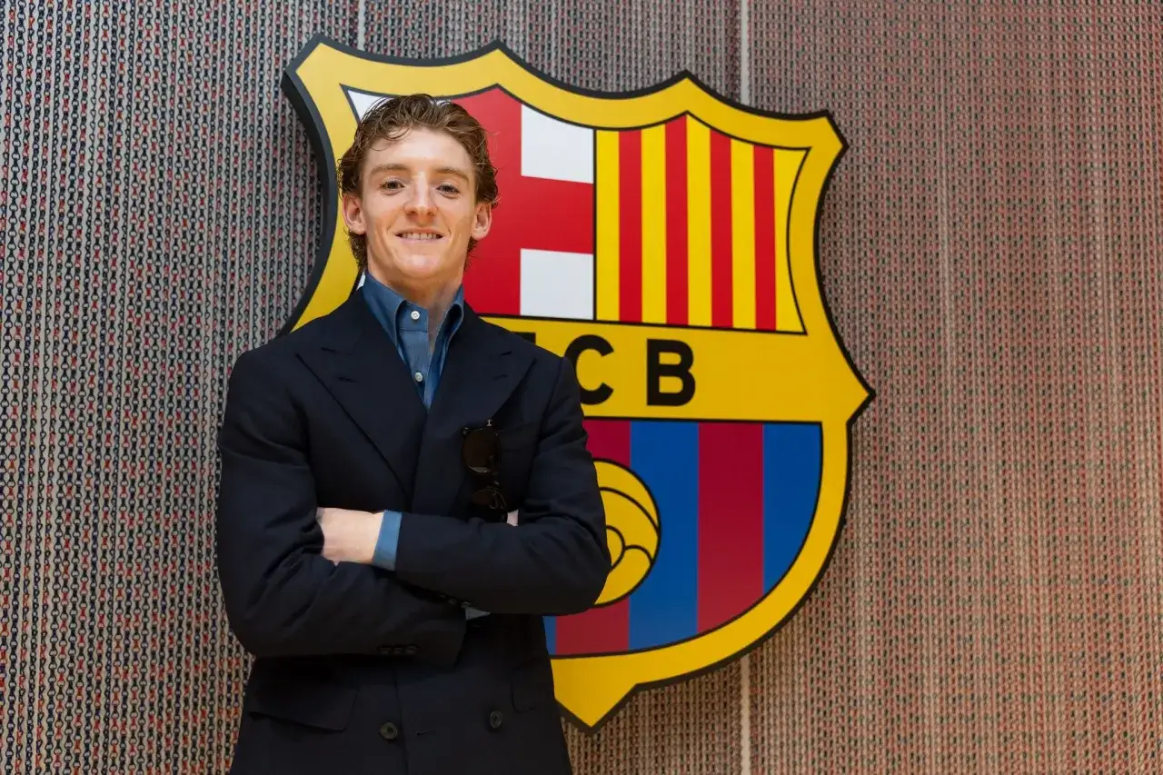 Uśmiechnięty Anthony Gordon w garniturze stoi przed herbem FC Barcelony.