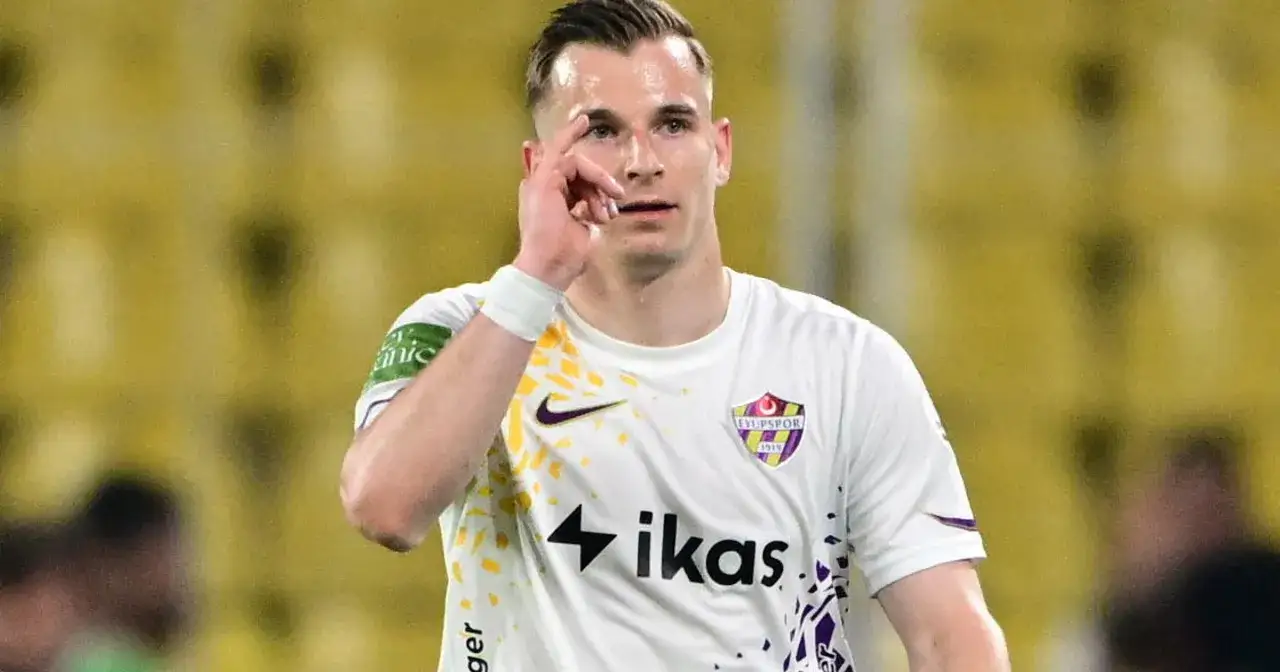 Piłkarz Mateusz Legowski w białej koszulce z herbem klubu i logo sponsora, gestykuluje palcem wskazującym przy skroni.