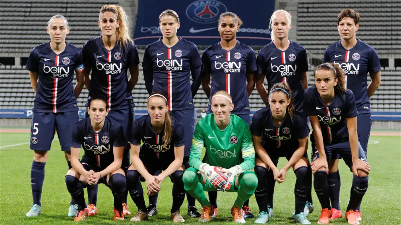 Drużyna PSG z Oliwia Szperkowską w składzie pozuje do zdjęcia. Piłkarki w granatowych strojach sportowych na zielonej murawie.