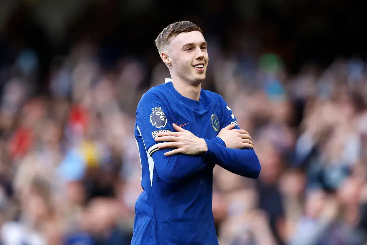 Cole Palmer w niebieskiej koszulce Chelsea z ramionami skrzyżowanymi na piersi, uśmiecha się do publiczności.