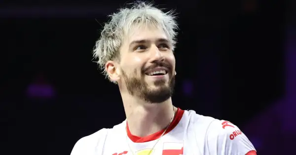 Uśmiechnięty siatkarz z blond włosami, jeden z tych, którzy wygrali Ligę Mistrzów, w białej koszulce z polską flagą.