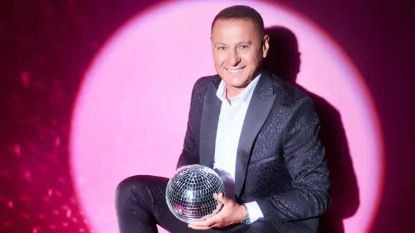 Piotr Świr Świerczewski w eleganckim garniturze trzyma błyszczącą kulę dyskotekową na tle różowego światła.