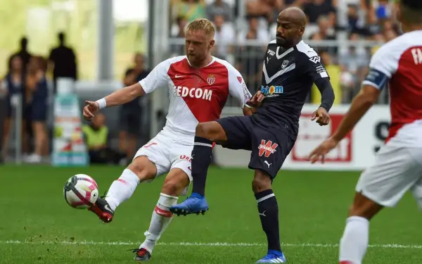 Piłkarz w białej koszulce z herbem **AS Monaco FC** walczy o piłkę z przeciwnikiem w granatowym stroju.