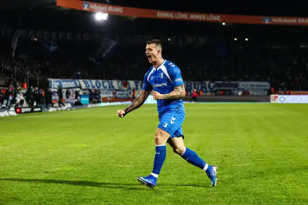 Mateusz Żukowski w niebieskim stroju świętuje bramkę na zielonej murawie stadionu, z trybunami pełnymi kibiców w tle.