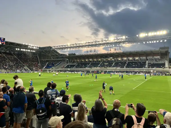 Widownia na stadionie przed meczem FC Bologna. Piłkarze rozgrzewają się na zielonej murawie, a kibice z flagami i aparatami obserwują przygotowania.