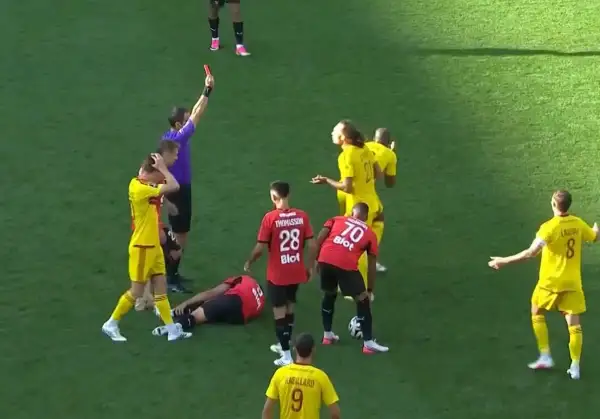 Sędzia pokazuje czerwoną kartkę w trakcie meczu Stade Rennais. Piłkarz leży na boisku, inni zawodnicy reagują.